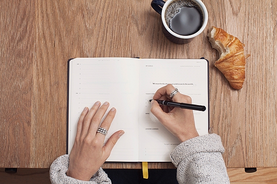 Tasse Kaffee, ein Croissant und ein Notizbuch liegen auf einem Tisch. Zwei Hände tragen etwas in das Notizbuch ein.