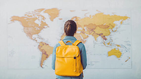 Eine junge Frau mit einen gelben Rucksack steht vor einer Landkarte, die die Welt zeigt