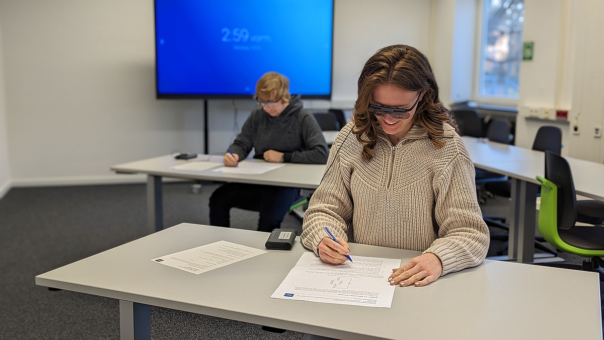 In dem Raum sind im Vordergrund zwei schräg an Einzelarbeitsplätzen hintereinander sitzende Personen mit VR-Brillen zu sehen. Sie füllen Arbeitsblätter aus. Im Hintergrund ist ein interaktives Smartboard und ein Fenster zu erkennen.