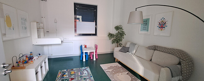 Blick in den Elternkindraum: rechts an der Wand befindet sich ein Sofa, links ein Wickeltisch, daneben mehrere Spielmöglichkeiten und ein kleiner Tisch