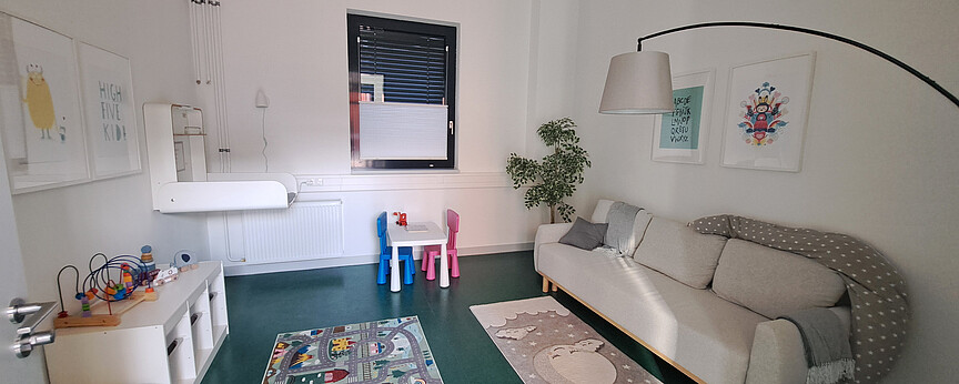 Blick in den Elternkindraum: rechts an der Wand befindet sich ein Sofa, links ein Wickeltisch, daneben mehrere Spielmöglichkeiten und ein kleiner Tisch